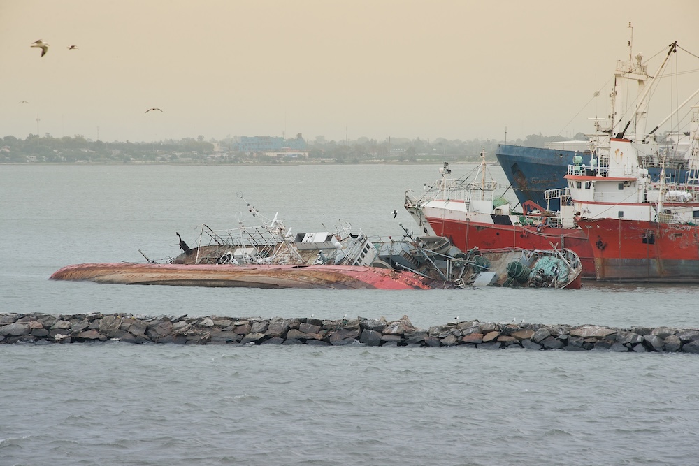 Capsized red and white ships partially submerged near a rocky sea barrier with seabirds in flight